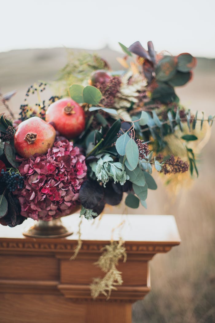 Vibrant floral arrangement featuring pomegranates and hydrangeas in rustic outdoor setting.