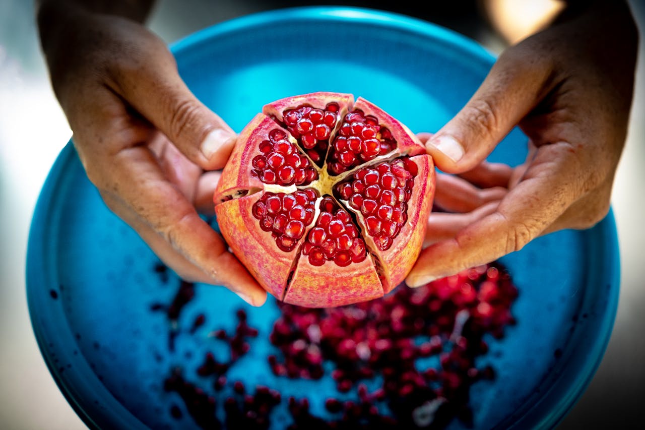 Close-up of a fresh juicy pomegranate being held over a blue bowl, revealing its vibrant red seeds.