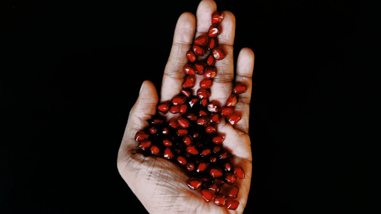 A close-up view of a hand holding vibrant pomegranate seeds against a dark background.